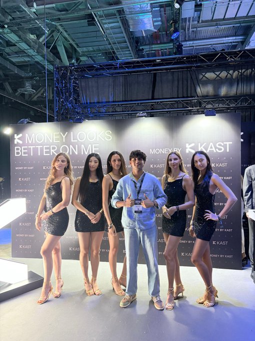 A group of six people poses at a 'MONEY BY KAST' event; five young women in matching sparkly black dresses flank a young man in a light blue shirt and jeans holding an award.