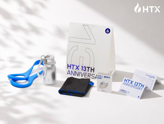 An HTX 13th Anniversary promotional display featuring a paper bag, silver water bottle, blue strap, black pouch, and promotional cards arranged on a white surface.