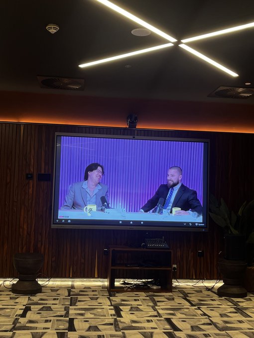 A large wall-mounted TV in a room with patterned carpet and wooden paneling displays a video podcast or interview featuring two men in suits sitting at a table with mugs and microphones.