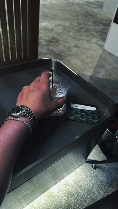 A hand adorned with a silver watch and bracelet reaches for a clear plastic cup of water with a straw on a dark tray, alongside a green card holder.