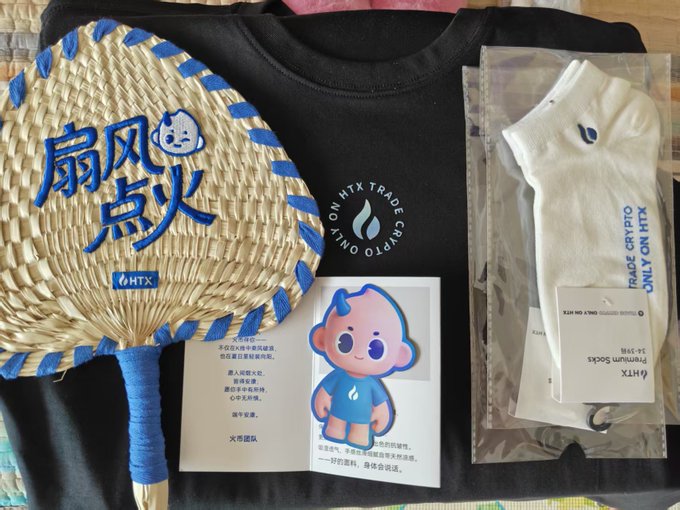 The image shows a Dragon Boat Festival gift box from HTX (Huobi) exchange, featuring a black t-shirt with "HTX INTO CRYPTO" branding, a handwoven fan with "扇风点火" (stir up trouble) text and the HTX logo, a Huobi "Huobao" mascot sticker with Chinese poetry, a plush toy in packaging labeled "TRADE CRYPTO ONLY ON HTX," and socks branded with the HTX name—all corresponding to the user's appreciation post about receiving quality merchandise from the exchange, particularly highlighting the liquid cotton texture of the seemingly plain t-shirt and the "fierce-looking" Huobao mascot sticker.