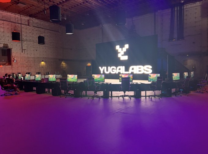 An interior photograph of a Yuga Labs event space featuring a large illuminated 'YUGA LABS' logo on a stage, with rows of computer monitors arranged on tables lit with purple ambient lighting.