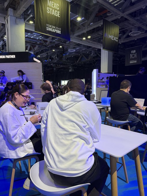 A bustling convention floor scene at the MEXC Token2049 Singapore event, showing attendees seated at tables working on laptops and phones under hanging 'MEXC Stage' banners.