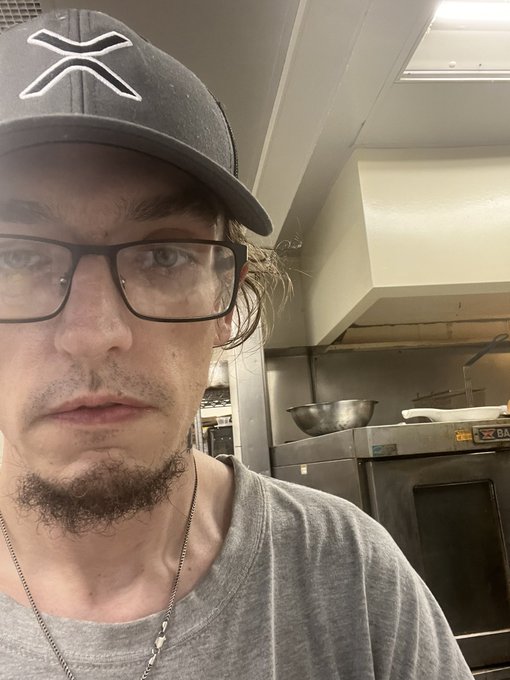 A selfie of a man with glasses, a goatee, and a gray cap with an 'X' logo, standing in what appears to be a commercial kitchen with a metal bowl and oven visible behind him.