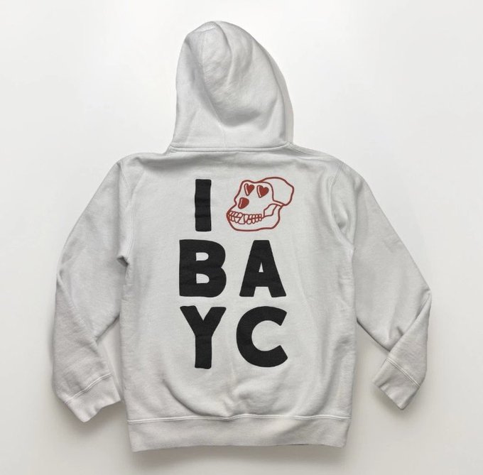 A light gray hoodie displayed on a white surface features the text "I ❤️ BAYC" in bold black lettering, with a red heart-eyed Bored Ape Yacht Club skull logo replacing the heart symbol. The image showcases the BAYC-themed apparel mentioned in the post, representing the new sweater the user acquired for their wife to celebrate their one-year "apeversary" anniversary.