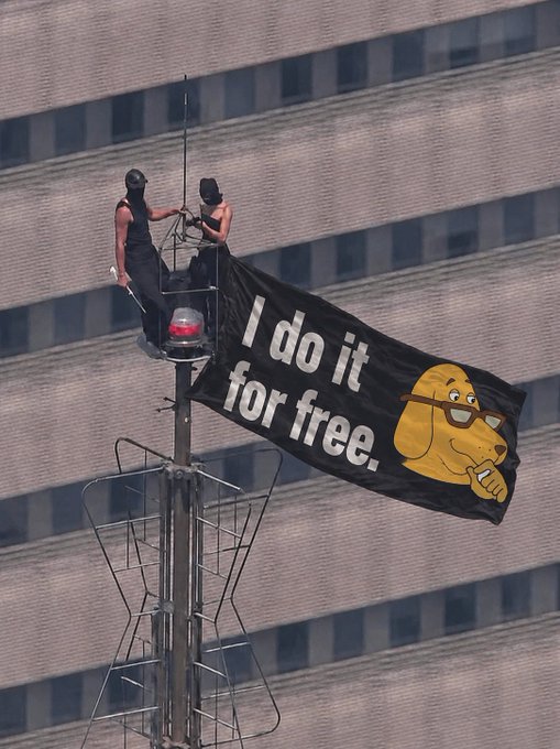 Two individuals in black attire and masks are suspended on a tall structure against a city building, displaying a black banner that reads 'I do it for free.' featuring an illustration of a dog wearing glasses.