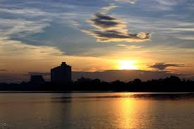 The image features a beautiful sunset over a body of water, with the sun setting behind some buildings or structures. The sky is filled with warm hues as the sun sets, creating a serene atmosphere. The scene captures the essence of a peaceful evening by the lake, making it an appealing and picturesque view for viewers to appreciate nature's beauty.