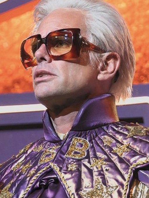 A close-up photograph of a man with white hair wearing large orange sunglasses and an ornate purple and gold jacket featuring a large 'B' on the chest.
