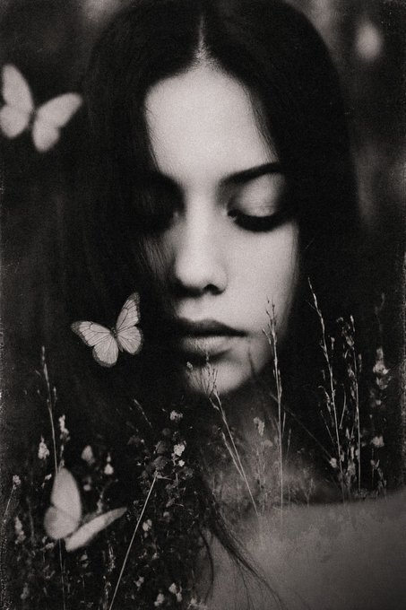 A black-and-white portrait of a young woman with closed eyes, surrounded by delicate wildflowers and accompanied by two white butterflies.