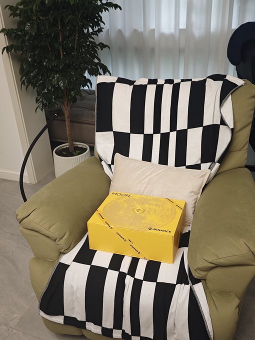 An olive green armchair draped with a black and white striped blanket, holding a white pillow and a yellow box labeled 'NOON' with the Binance logo.