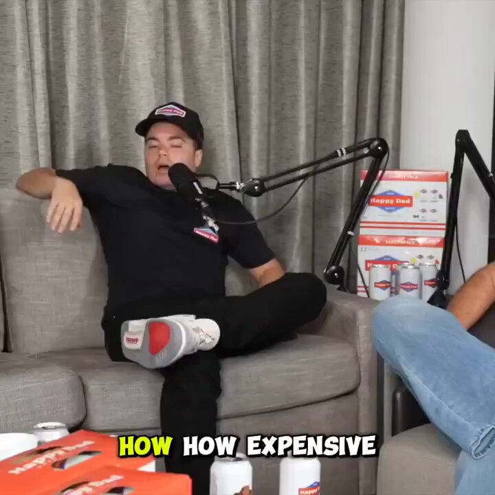 The image shows a man sitting on a couch with his legs crossed, wearing a baseball cap and black shirt. There are several cans of soda scattered around him, including two cans that appear to be beer cans. The background features a window with curtains, suggesting an indoor setting. The caption reads "How expensive" in the bottom right corner, which is likely related to the image's context or the post it belongs to.