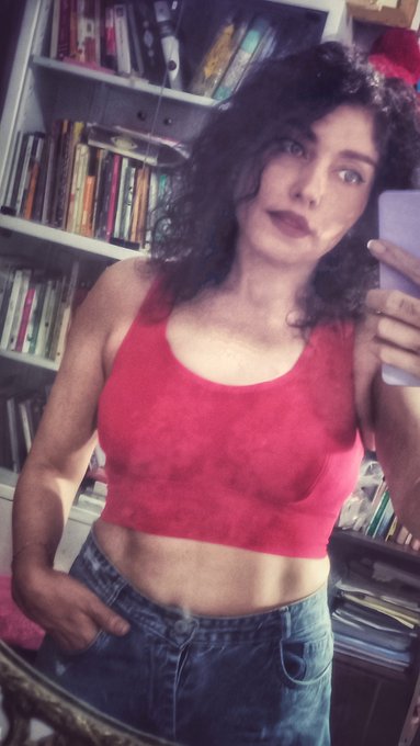 A woman with dark curly hair wearing a red cropped tank top and denim shorts poses for a mirror selfie in front of a bookshelf, holding a phone. The image complements the post promoting her "Crimson Veil" NFT collection (1/4 edition, 100 XTZ) on objkt.com, with her red outfit visually matching the artwork's crimson theme.