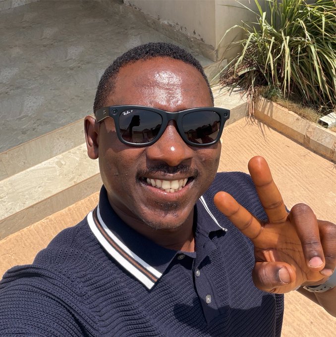 A smiling man wearing Ray-Ban sunglasses and a navy polo shirt flashes a peace sign, perfectly matching the cheerful "good morning, let's make it a good day" sentiment with his upbeat pose on sunlit stone steps.