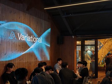 People sit at tables in a dimly lit venue with a glowing blue screen displaying the 'Variational' logo and a soundwave design.