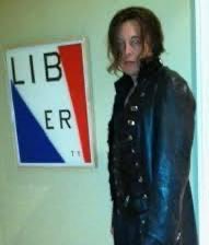 A person with long dark hair wearing a dark jacket stands next to a framed poster on a wall that reads 'LIB/ER' in red, white, and blue.
