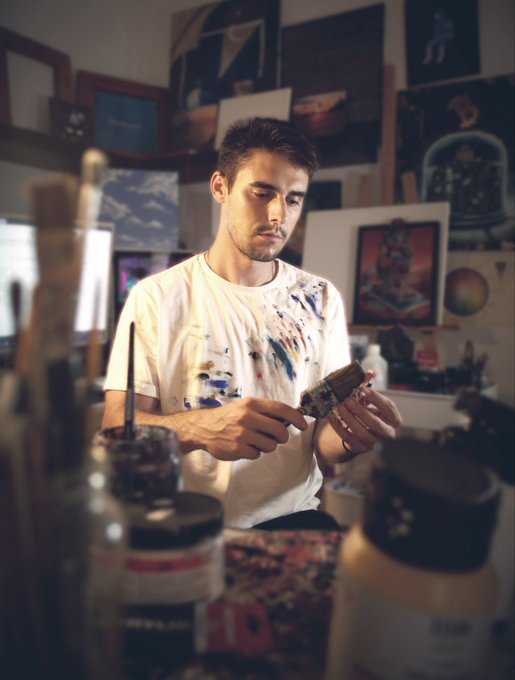 A young man in a paint-splattered white t-shirt stands in an art studio, examining a paintbrush among various artworks and supplies.