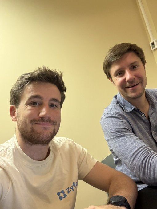 Two young men smile at the camera in a selfie-style photo against a plain beige wall; the man on the left wears a white t-shirt with 'Zyfo' visible on it.