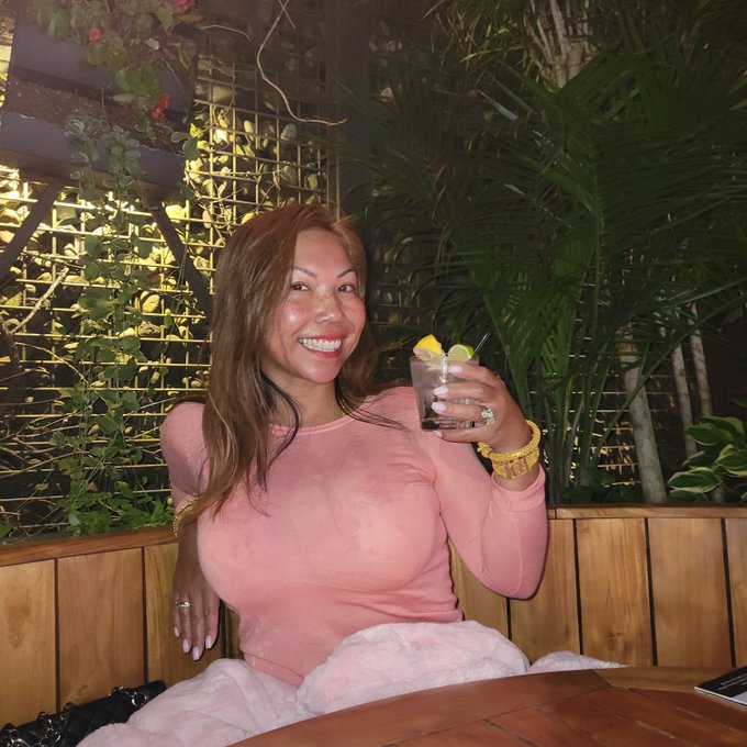 A woman with long brown hair wearing a pink long-sleeved top smiles while holding up a cocktail garnished with a lemon slice, seated at a wooden table in a plant-filled venue.