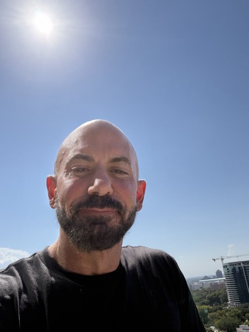 A bald man with a beard takes a selfie outdoors against a clear blue sky with bright sunlight, with a cityscape and construction crane visible in the background.