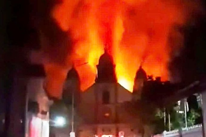 A nighttime photo of a domed building engulfed in massive orange flames and thick smoke, resembling a cathedral or church on fire.