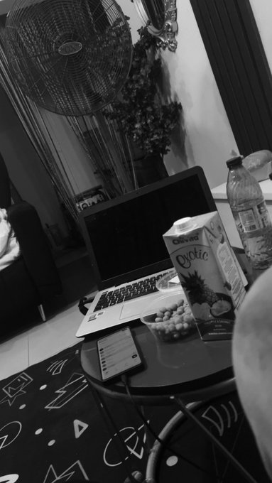 A black-and-white photo showing a laptop on a small table next to a carton labeled 'Exotic' and a bowl of snacks, with a standing fan in the background.