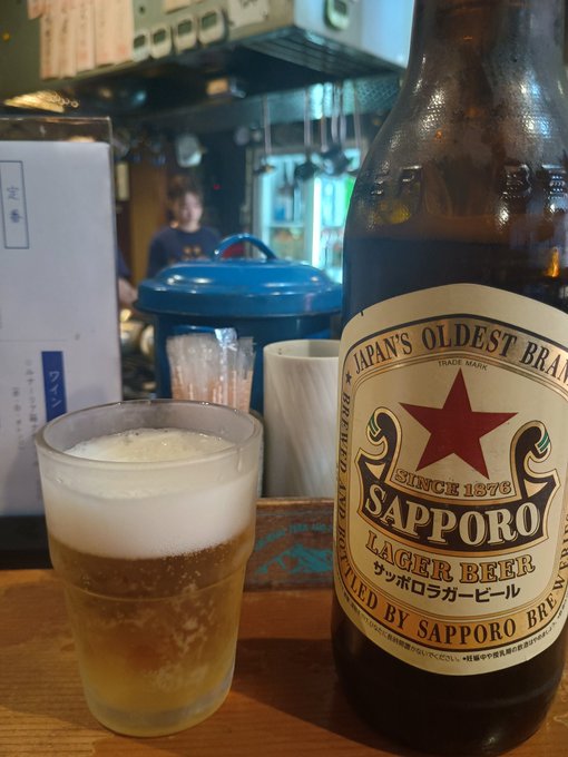 A glass of Sapporo lager beer filled with a frothy head sits beside its branded bottle on a wooden bar counter, with a Japanese izakaya interior visible in the background.