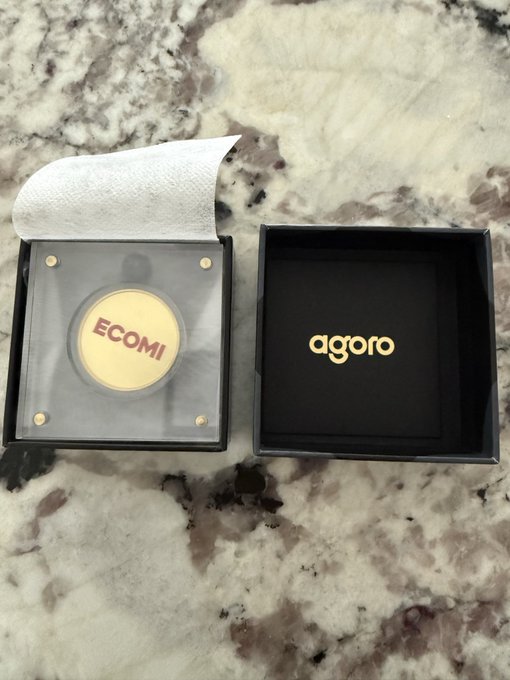 The image features a black box with the word "agoro" written on it, placed on top of a marble countertop. The box has a gold label that says "ecomi", indicating that it might be related to the cryptocurrency market or a product from the ecomi company.