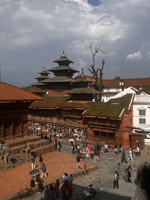 A bustling public square in a South Asian city (likely Kathmandu Durbar Square) featuring traditional pagoda-style temples with multi-tiered roofs, surrounded by red brick buildings and many pedestrians under a cloudy sky.
