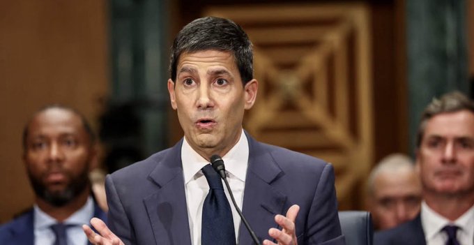 A man in a dark suit and tie speaks at a microphone, gesturing with his hands, with other people visible behind him.