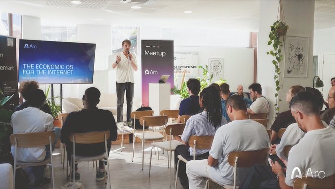 A presenter stands at the front of a room discussing "The Economic OS for the Internet" on a screen displaying the Arc logo, in front of an audience of around 20 people, with a "Meetup" banner visible in the background. The image captures the office presentation event where the speaker introduced the Arc vision ahead of the mainnet launch mentioned in the post.