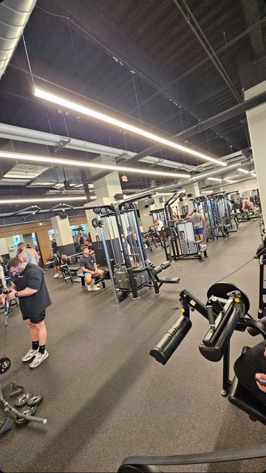 The image shows a large, well-equipped gym with multiple exercise machines such as treadmills, weight machines, and dumbbells. There are several people working out at various stations within the gym. One person can be seen wearing a black shirt while using one of the treadmills. The gym appears to be clean and organized, providing an ideal environment for fitness enthusiasts to work on their health and well-being.