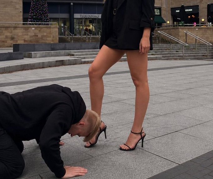 A photo of a man in a black hoodie kneeling on a stone plaza with his head bowed near the feet of a woman wearing a short black dress and high heels; a Christmas tree and storefronts are visible in the background.