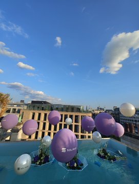 An outdoor scene showing purple and white spheres bearing the 'TAPBIT' logo floating above a pool on a rooftop, set against a city skyline and blue sky with clouds.