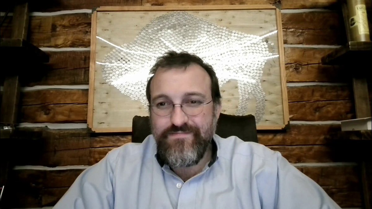 The image shows a man wearing glasses sitting at a desk, with a wooden wall behind him featuring a large metal art piece depicting a buffalo. The man appears to be looking directly into the camera while seated in an office chair. The image also contains text that reads "Cardano is about to shift from a research-first chain to a throughput-first beast", which suggests that there might be some sort of technological or business change happening related to Cardano, possibly involving its integration with Leios and Peras.