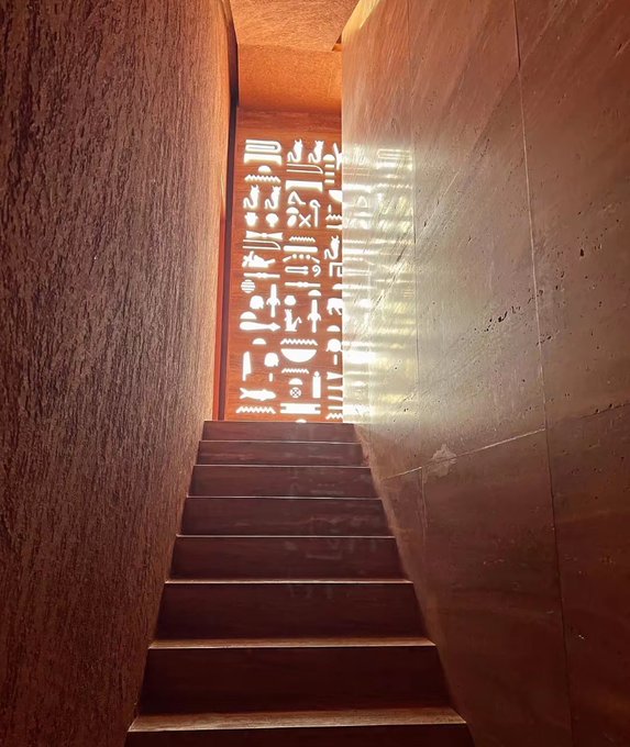 A wooden staircase ascends toward an illuminated wall displaying hieroglyphic-style symbols, creating a dramatic, tomb-like atmosphere. The image visually echoes the post's "lock in and go all in" message, symbolizing a deliberate climb toward opportunity in $GM CT as the trader prepares to hunt the next runner.