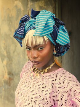 A young woman with striking blonde bangs wears an elaborate blue and teal striped headwrap, gold earrings, and a layered gold necklace, paired with a textured pink garment. The bold, fashionable styling embodies the "Solana Season" vibe, evoking vibrant confidence and contemporary cultural flair.