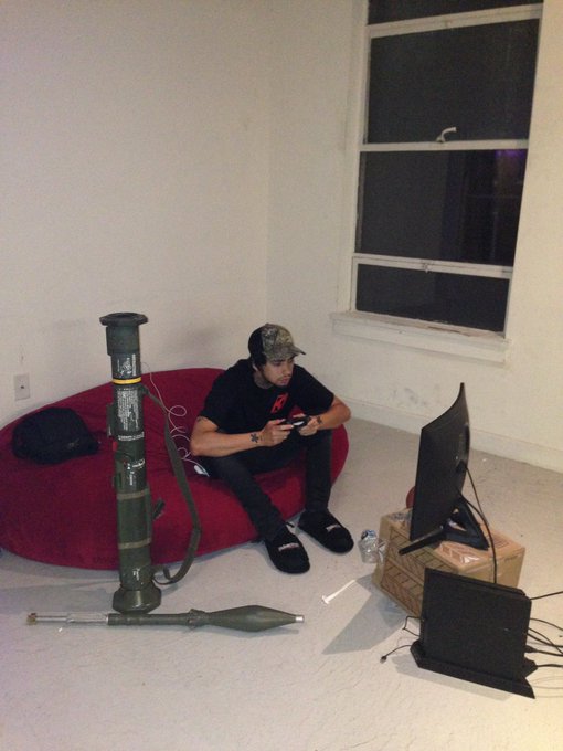 A young man in a black t-shirt and cap sits on a red beanbag chair playing a video game, with a missile-shaped object on the floor beside him.