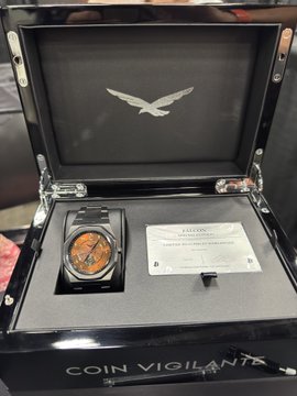 An open presentation case labeled 'COIN VIGILANTE' displaying an orange-faced luxury watch, a certificate of authenticity, and an eagle emblem.