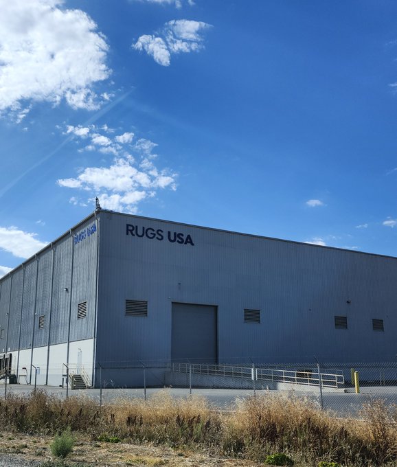 A large white industrial building labeled 'RUGS USA' stands behind a chain-link fence under a blue sky with scattered clouds.