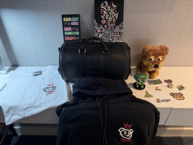 A collection of merchandise including a black duffle bag, a white t-shirt and black hoodie with a pixelated logo, a stuffed dog toy, and various stickers displayed on a desk.