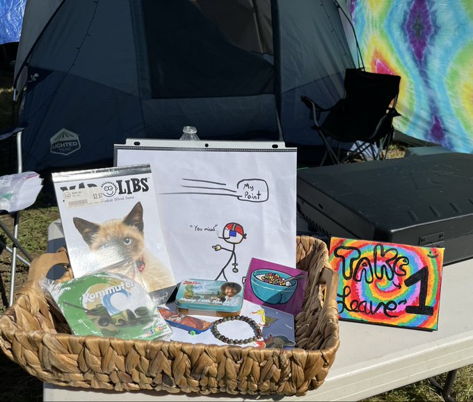 A woven basket filled with assorted thrift store finds—including a "Cat Hats LIBS" card game, "Rampage" game, a ceramic bowl, beaded jewelry, and a colorful tie-dye "Thanks 4 Leave" sign—sits on a table at a campsite, fittingly illustrating the "one man's trash is another man's treasure" sentiment as discarded or donated items are repurposed for camping entertainment.