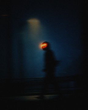 A blurry, motion-streaked silhouette of a person walks past a wall, illuminated by a single beam of light in an otherwise dark environment, creating an eerie atmosphere.