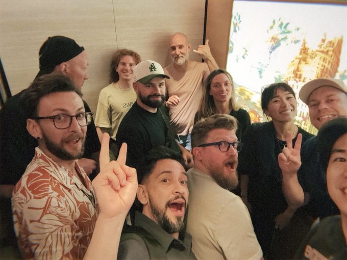 A group photo of about eight people gathered closely together indoors, smiling and posing with peace signs, with what appears to be classical artwork visible on a screen in the background.