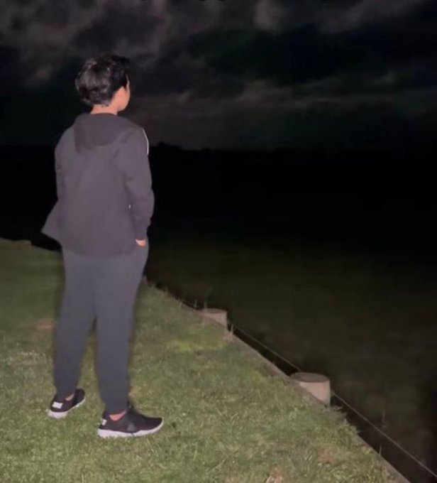 A young man in a dark hoodie and sweatpants stands alone on a grassy hillside at night, gazing out into the darkness with his hands in his pockets. The solitary, contemplative scene visually echoes the post's introspective question about whether 5 million impressions is an unattainable myth.