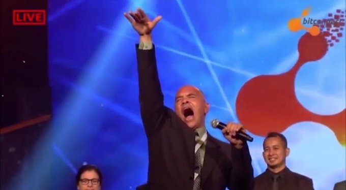 A live broadcast screenshot showing a bald man in a suit passionately speaking or singing into a microphone with his arm raised, with a "LIVE" badge and a Ripple-style logo visible in the background.