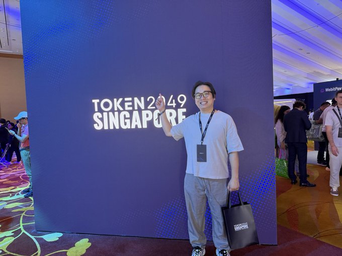 A man wearing glasses and a lanyard poses cheerfully in front of a large blue "TOKEN 2049 SINGAPORE" backdrop at the crypto conference, holding a branded event tote bag while flashing a peace sign. The image aligns with his post about attending #token2049 in Singapore, where he's networking and inviting others to connect.