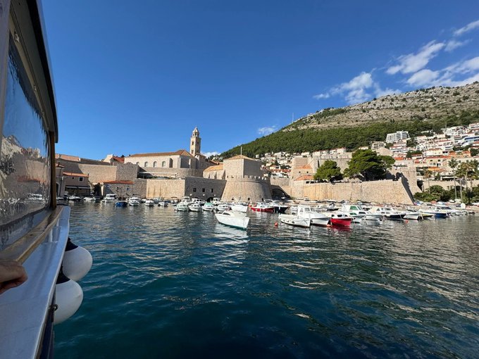 A scenic coastal view from a boat showing historic stone walls, a church bell tower, and boats docked in turquoise water with mountains in the background.