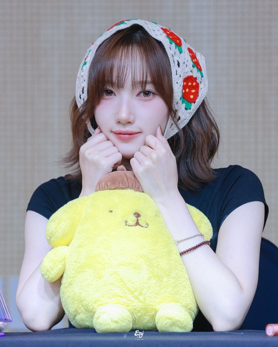 A young woman wearing a knitted strawberry-patterned bonnet smiles while resting her chin on her hands, holding a yellow plush toy resembling Pompompurin.