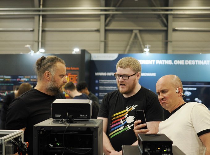 The image shows three men standing around a display table, with one man holding up his phone while looking at another man's cell phone. The third man is also looking at the phones. They appear to be discussing or comparing their devices. In the background, there are other people present in the room, and a sign can be seen hanging on the wall that reads "Recap of the first day at @BTCPrague: This stuff is soo freakin cool!".