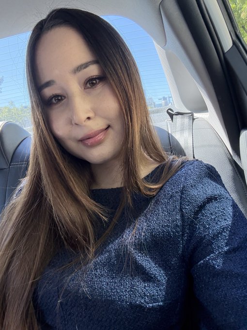 A selfie of a woman with long brown hair wearing a dark blue textured sweater, taken inside a vehicle with a sunny outdoor view visible through the window.